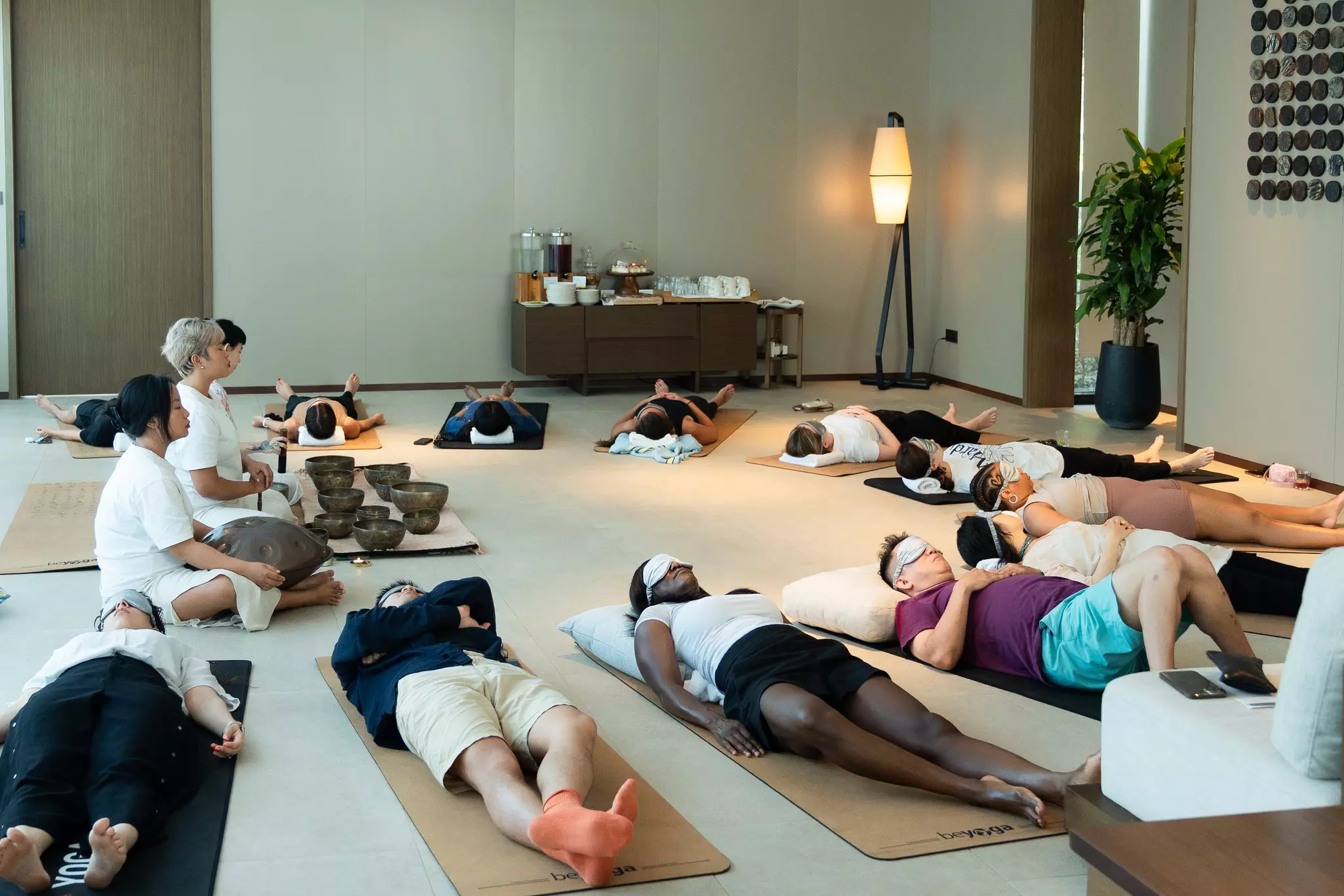 People in eye masks lying on mats in a half circle around three people who are sitting cross-legged with various bowls in front of them