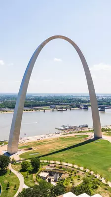 An aerial view of a monument in the shape of a monumental parabola, by a river in a city.