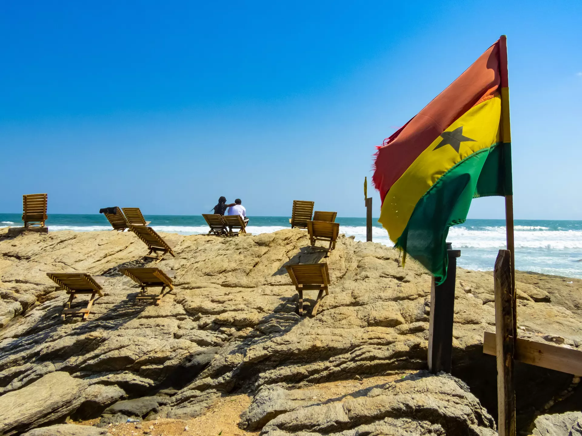 A yellow, red and green flag flies in the wind on a rocky outcrop near a beach where two people are sitting.