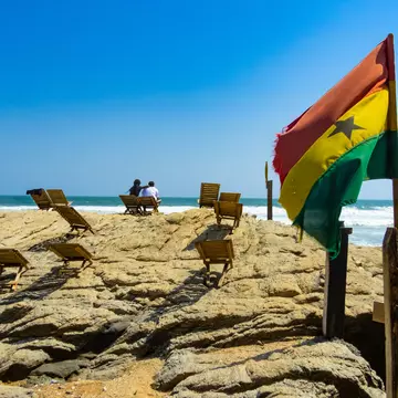 A yellow, red and green flag flies in the wind on a rocky outcrop near a beach where two people are sitting.