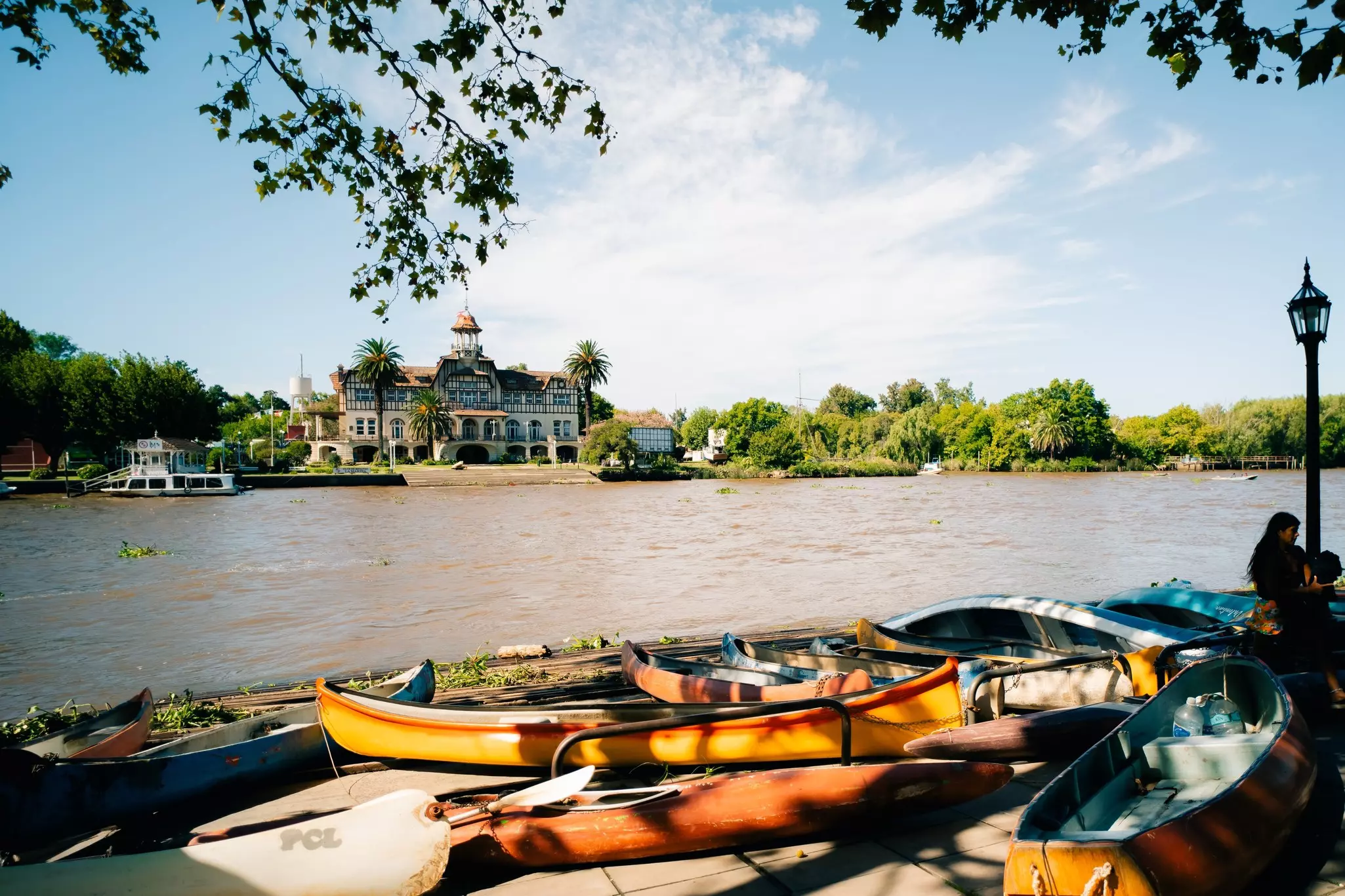 Small wooden canoes docked beside a wide river with a large grand building on the opposite side.