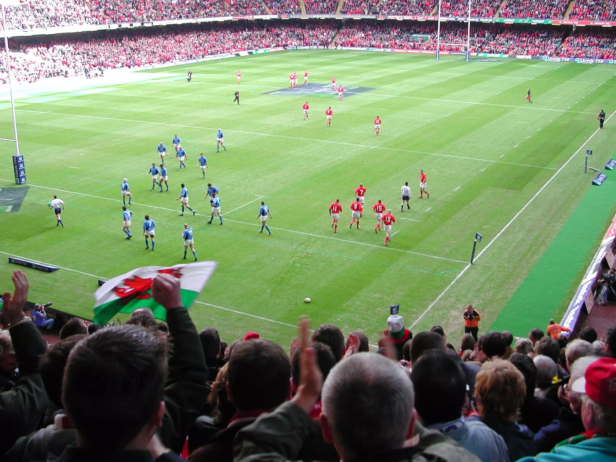 People wave flags and cheer in the bleachers while watching a rugby match in a huge stadium.