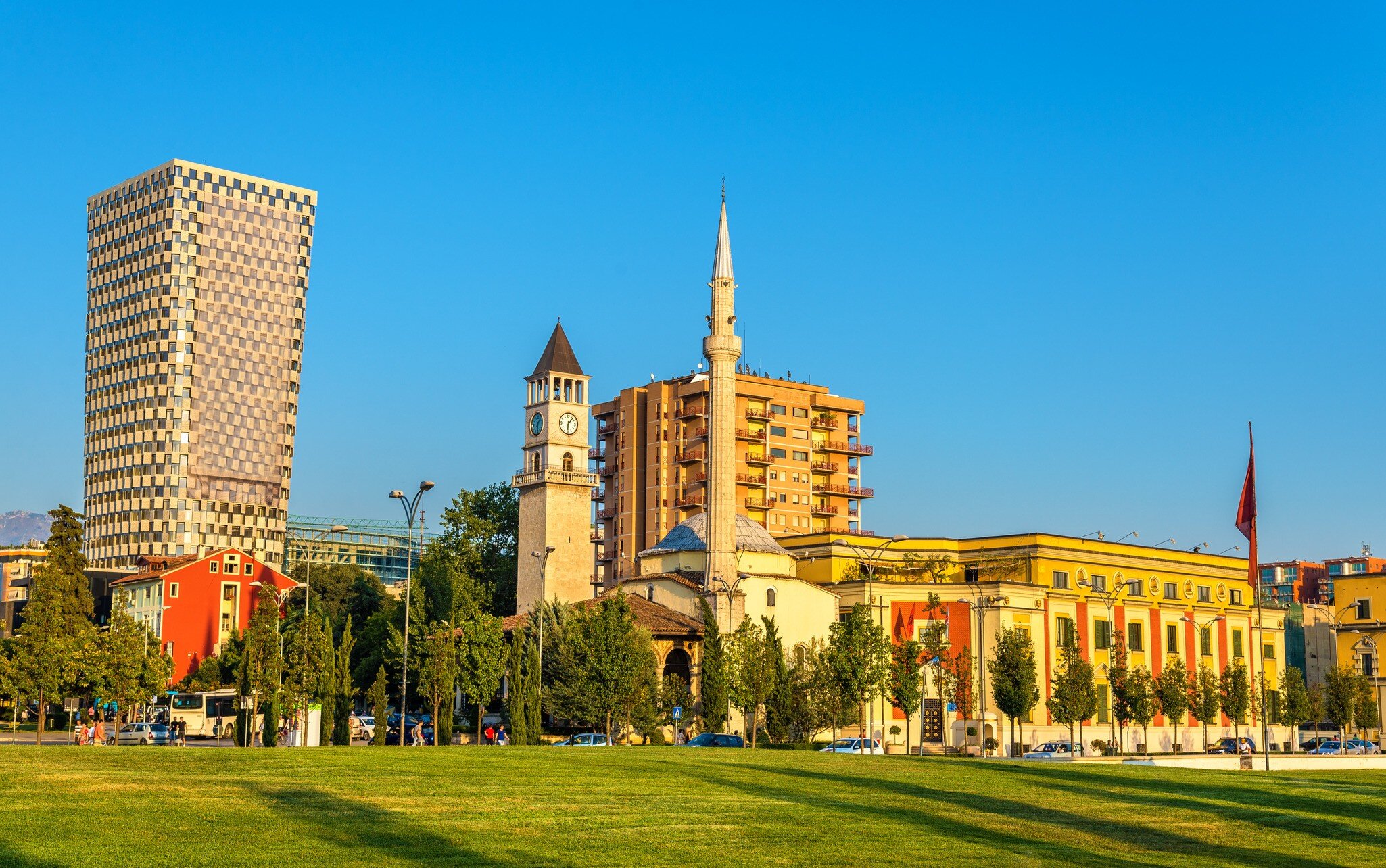 Slender white minaret and small domed mosque next to a stone clock tower, with a lawn in front and tower blocks behind