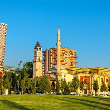 Slender white minaret and small domed mosque next to a stone clock tower, with a lawn in front and tower blocks behind