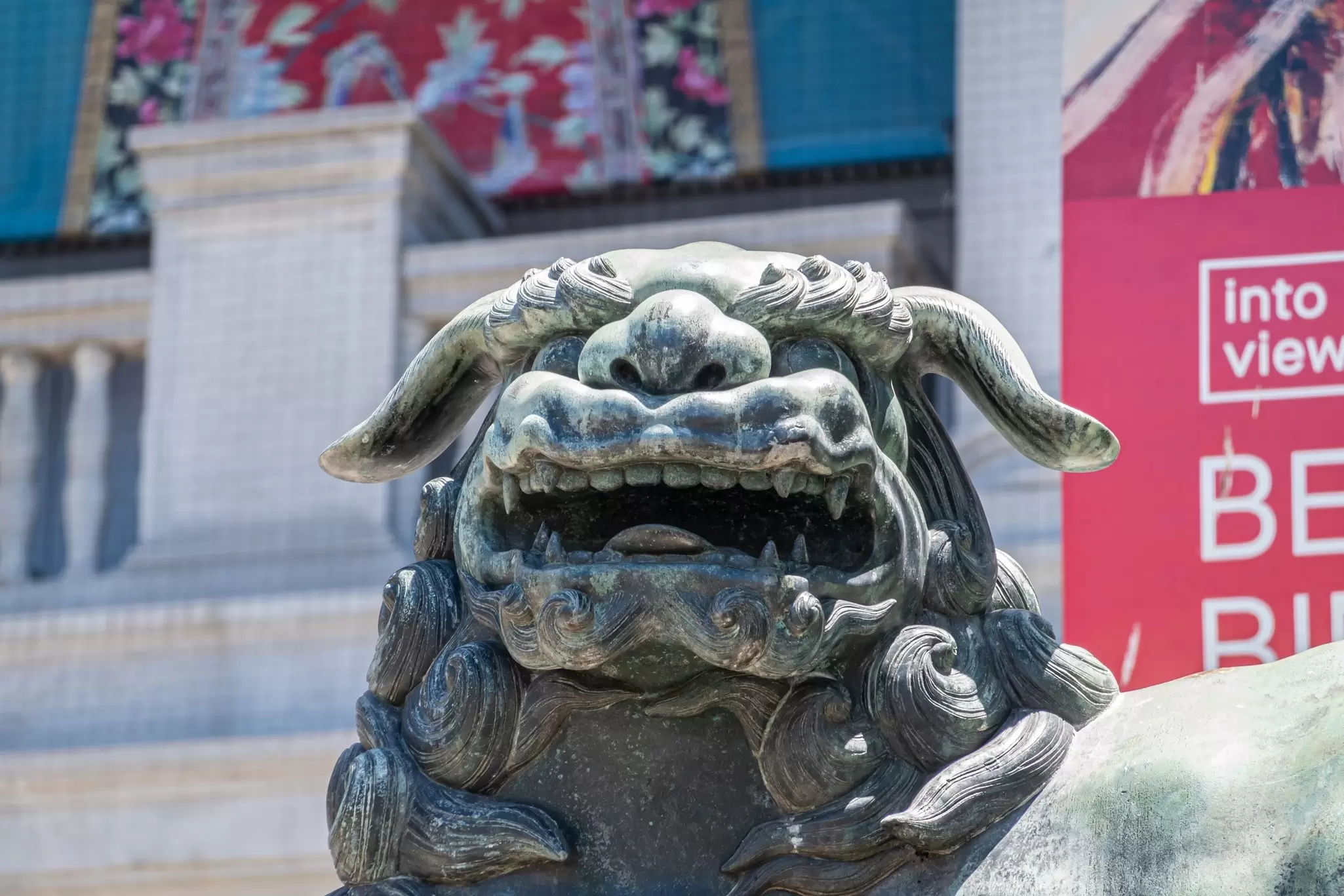 Bronze statue of a lion, facial closeup, in front of Asian Art Museum facade with its colorful banners promoting its treasures.