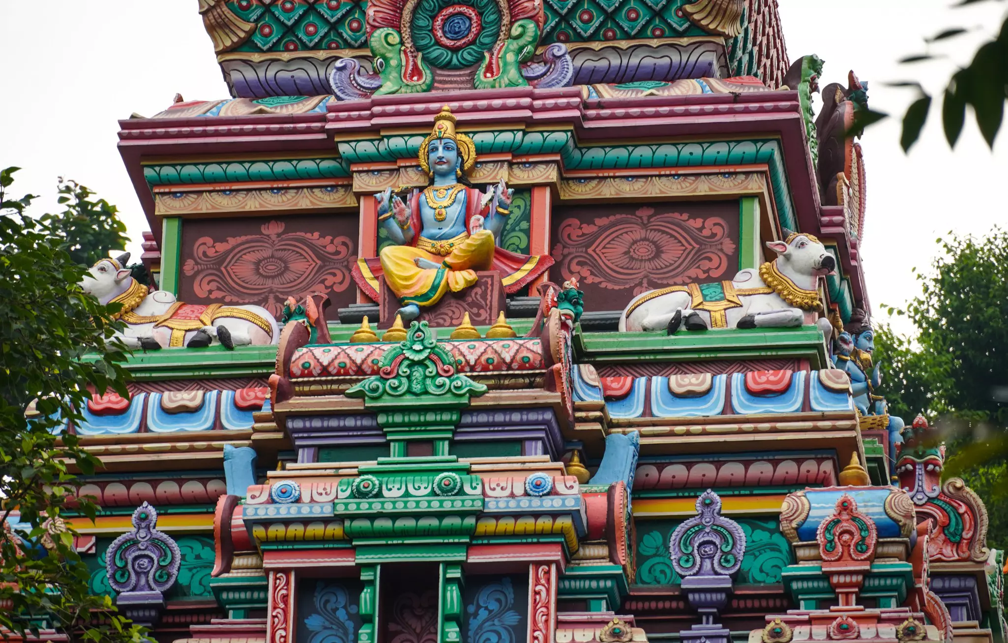 Top of colorful, ornate temple depicting Lord Shiva next to two bulls on an overcast day.