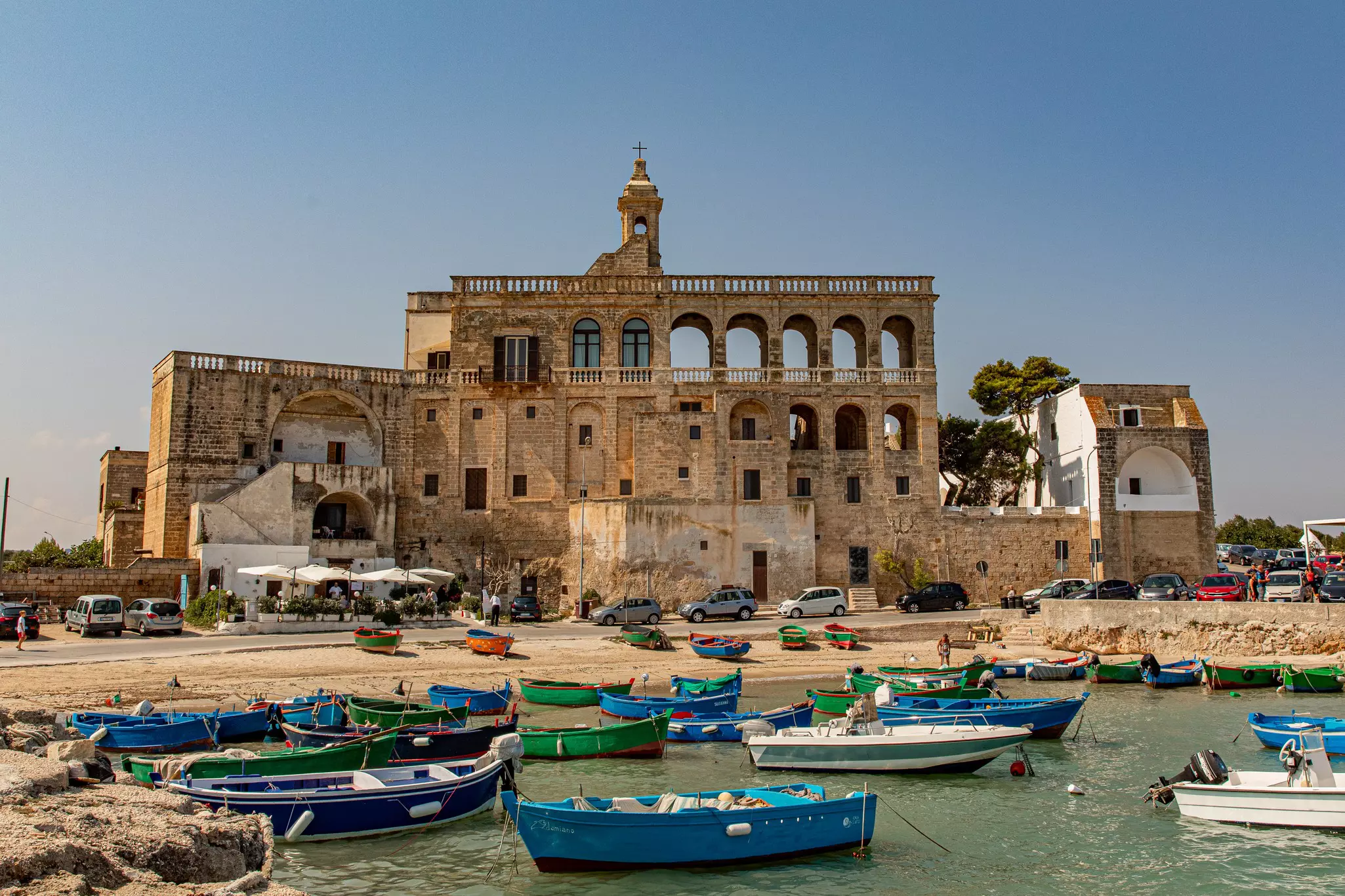 A tan stone abbey in front of a small sandy beach with small boats in the water.