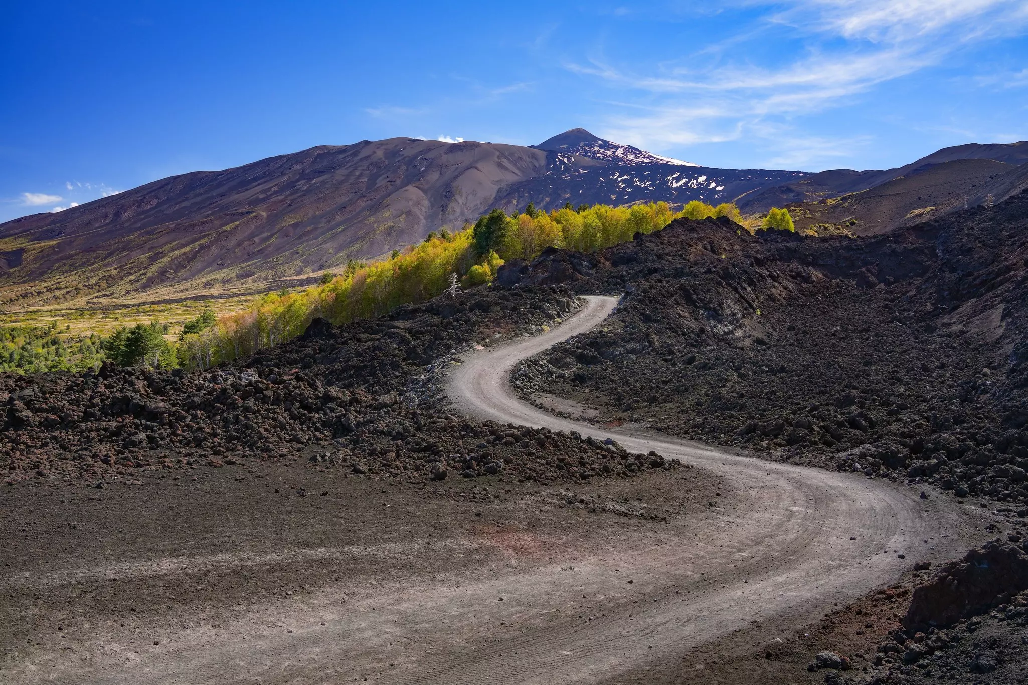 A windy dirt road on a volcano wends past volcano craters and vegetation.