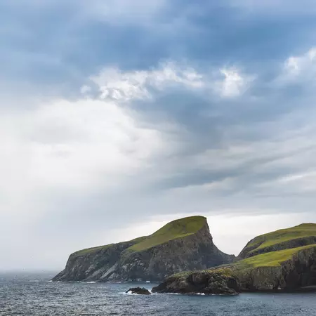 Green coastal cliffs set in dark, choppy waters, with cloudy gray skies overhead. 
