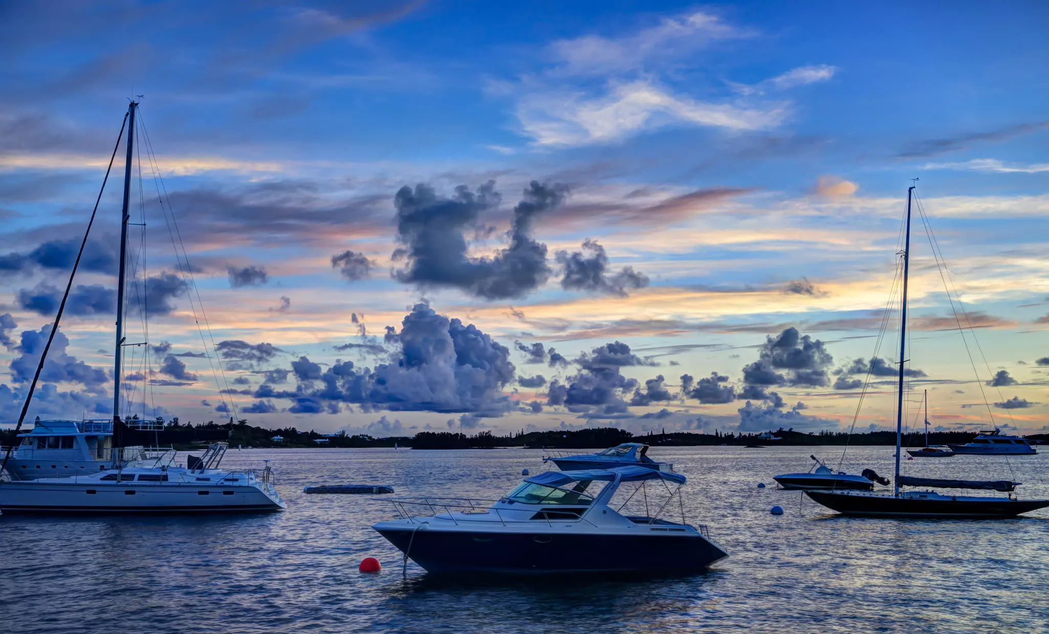 Small boats moored in a harbor below a sky of blue clouds as the sun sets.