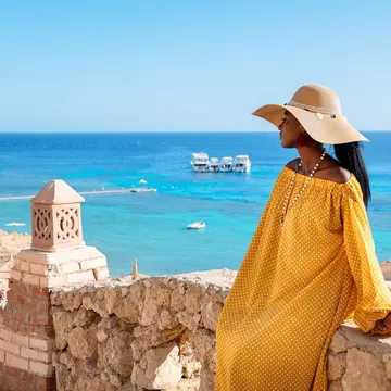 A woman wearing a yellow dress and a sunhat stands at an overlook above a sandy beach and turquoise sea in Sharm El Sheikh, Egypt