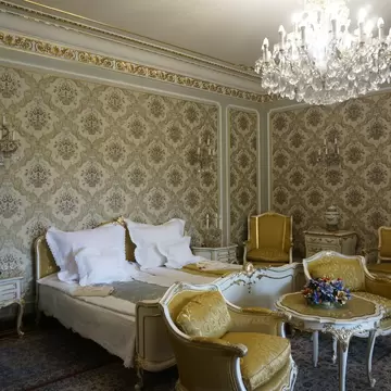 An ornate bedroom with damask-papered walls, a crystal chandelier, a white-and-gold bed and yellow silk armchairs.