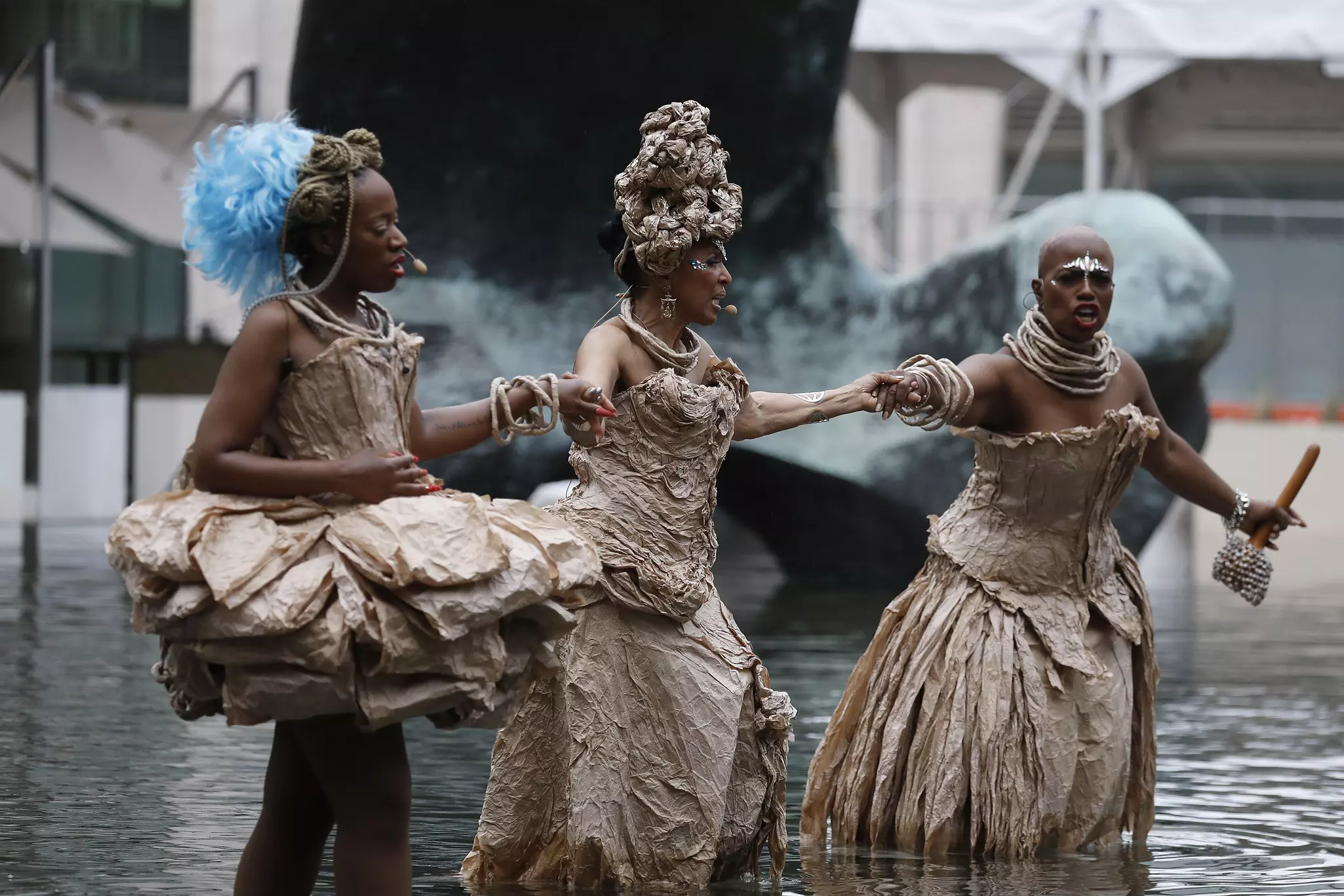 Three women in beige costumes stand in the water during a performance outside Lincoln Center