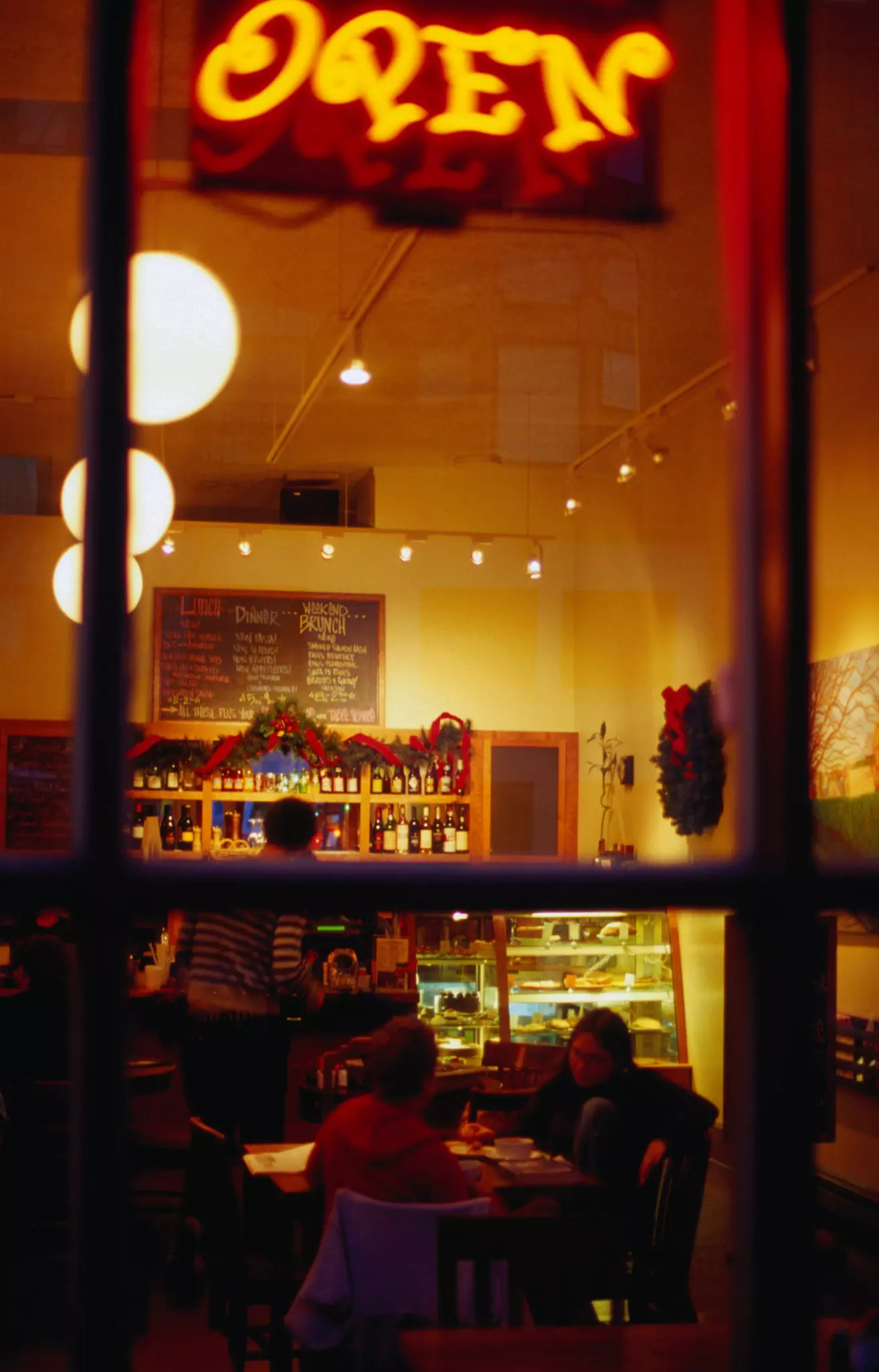 A view through a window of a dimly lit coffeehouse. Two people are sitting at a table, one facing the window and one whose back is to toward the window.