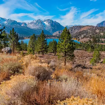 Blue lake among Jeffrey pines beneath snow-dusted granite peaks, golden rabbitbrush and dry grasses in the foreground