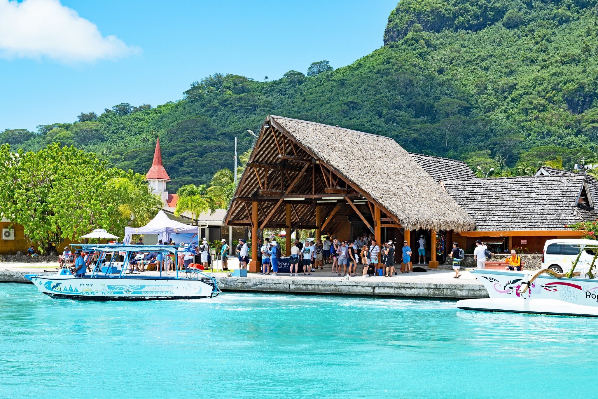 Crowd beneath a large palm-thatched shelter at a quay, a boat on turquoise water and a church spire against green hills
