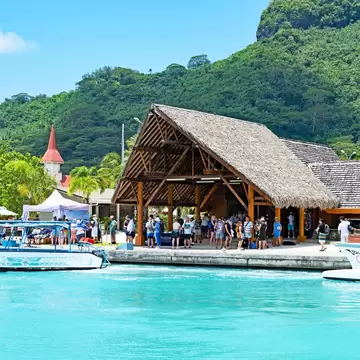 Crowd beneath a large palm-thatched shelter at a quay, a boat on turquoise water and a church spire against green hills