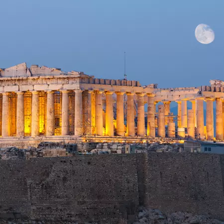The lit-up structure of the Parthenon at night, with the moon in the background. 