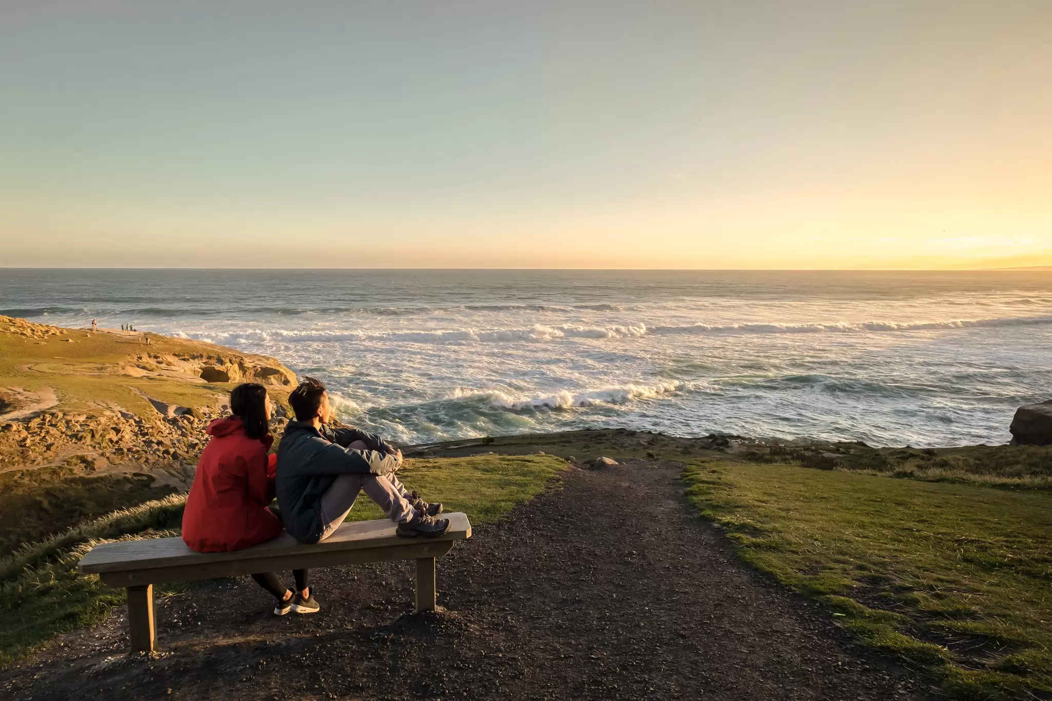 Couple sitting on a bench at the end of a path looking out over a rocky coastline and waves at sunset