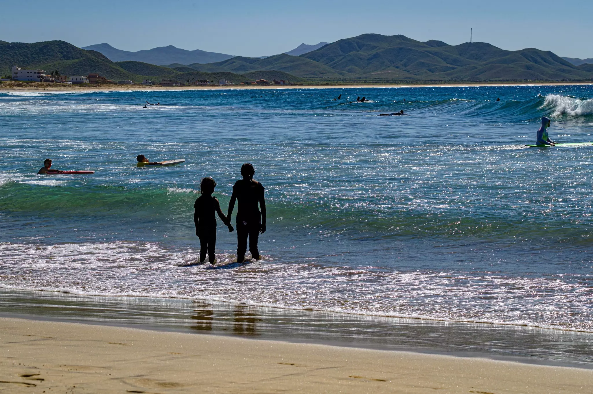 Two people hold hands while wading into water from a beach; surfers are paddling in the water.