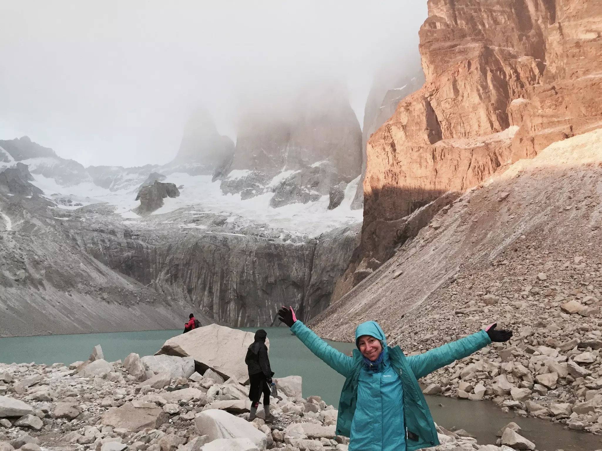 A hiker poses with arms in the air in front of a glacial lake where the sky is obscured by low-hanging clouds