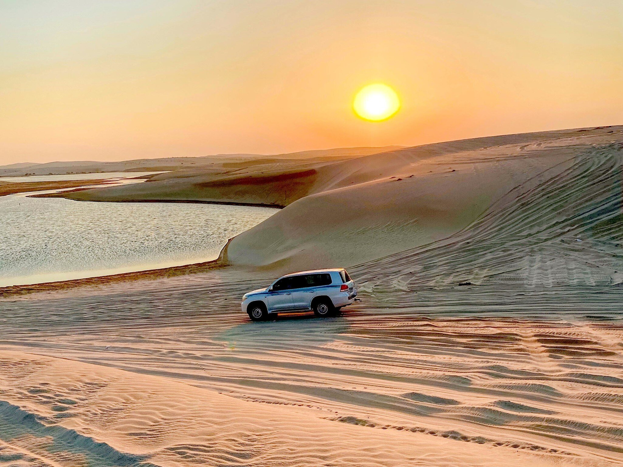 White SUV parked on tyre-tracked sand dunes beside a calm lagoon as the sun sets in a hazy orange sky