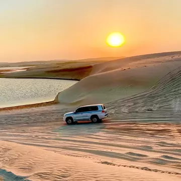 White SUV parked on tyre-tracked sand dunes beside a calm lagoon as the sun sets in a hazy orange sky