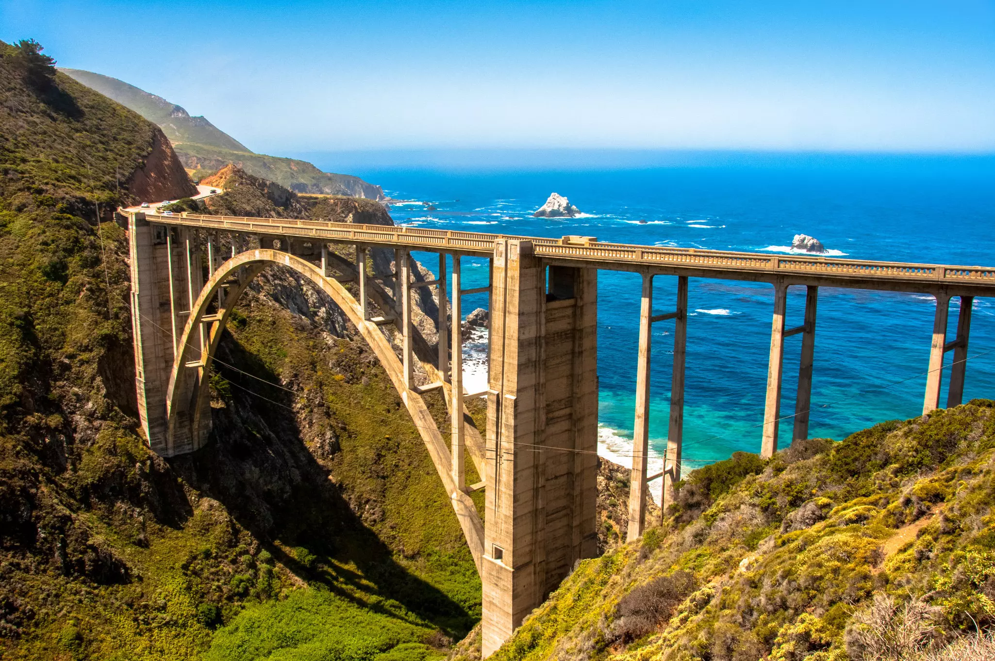 A massive single-span bridge made of concrete curves around a small ravine on the coast of California near Big Sur