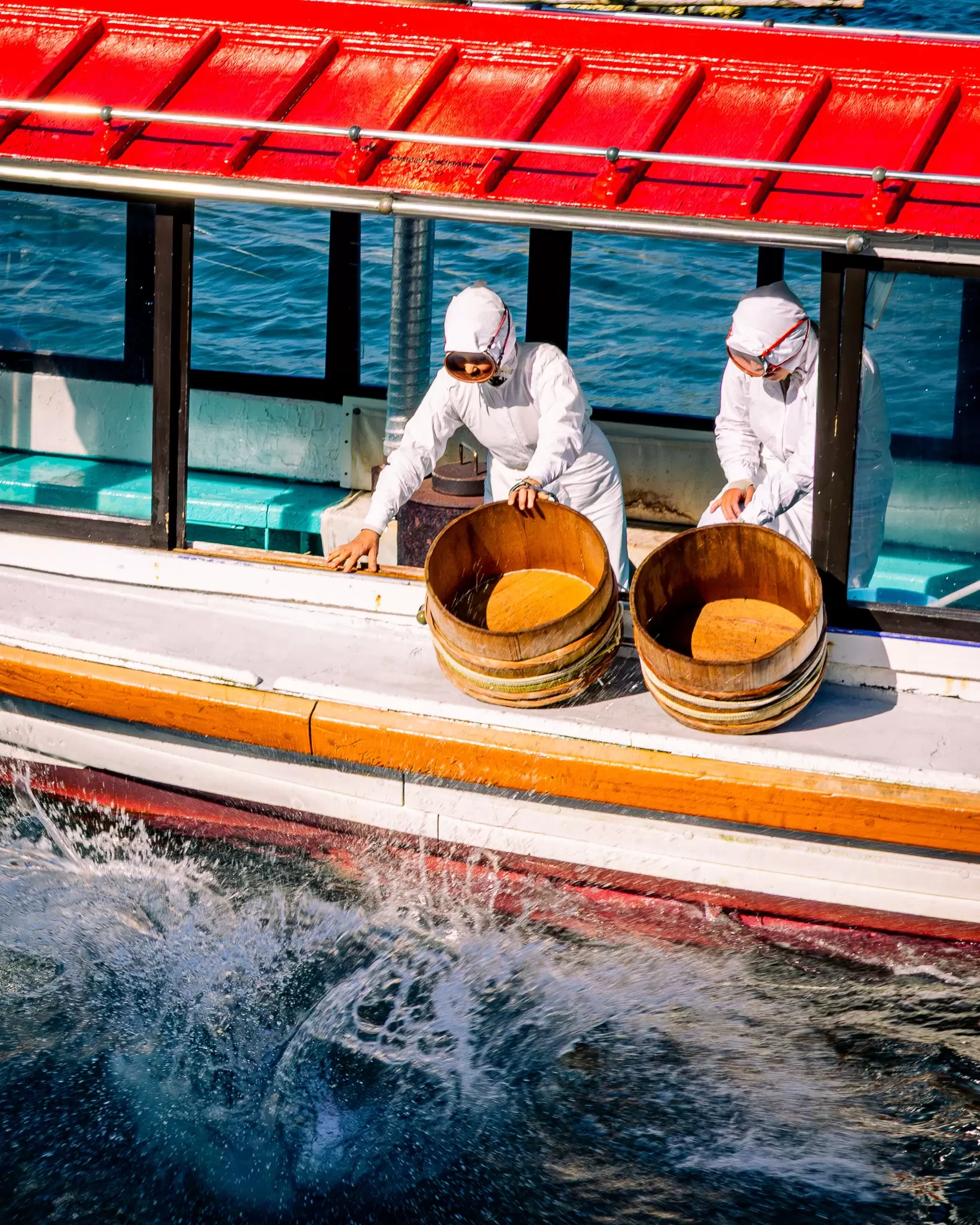Divers in all-white uniforms prepare to jump into the ocean off a small boat, holding onto wooden buckets. 
