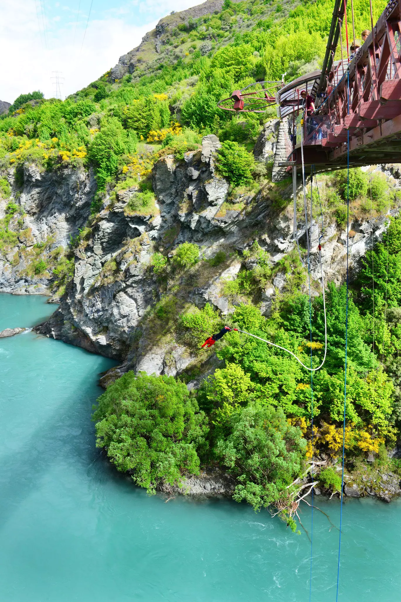 A person, with their arms out above their head, attached to a cord by their ankles, plummets towards a blue river from a bridge.