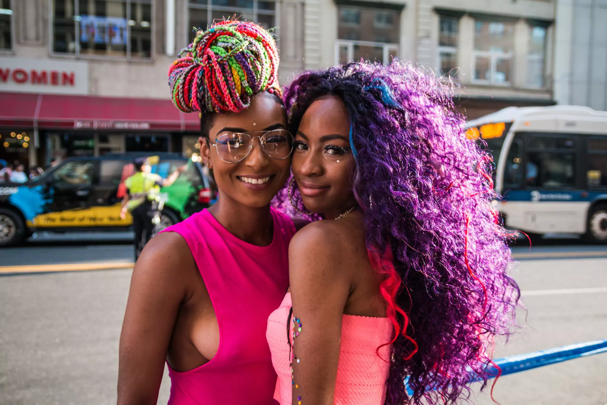 Two women wearing pick tops and with colorful hairstyles are pictured in a city street.