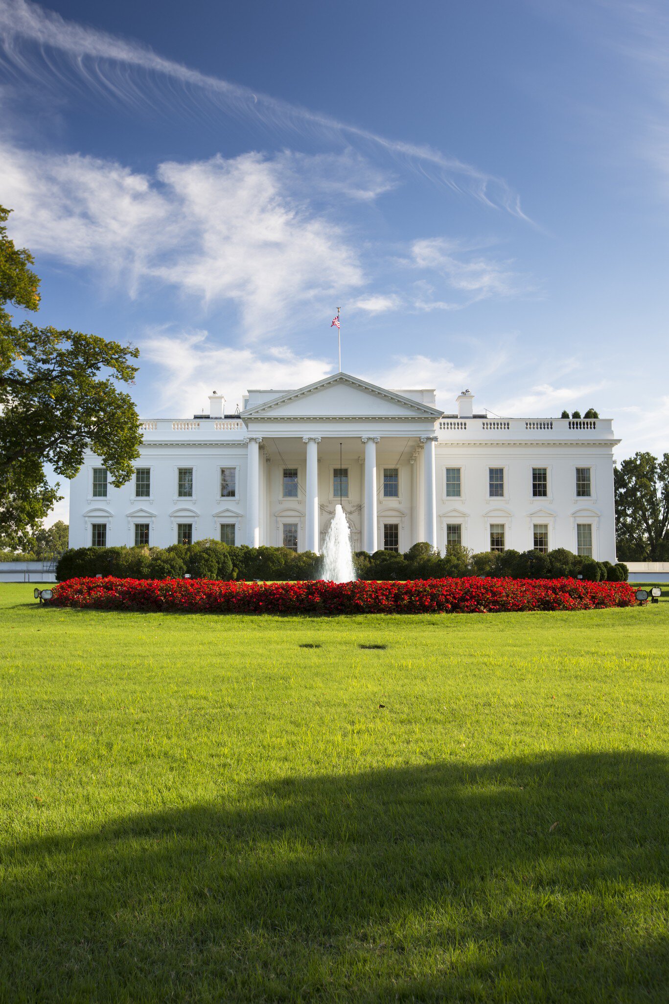 Columned portico of a white mansion behind a fountain and a curved bed of red flowers on a sunlit green lawn.