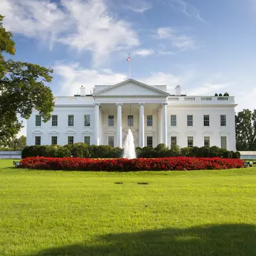 Columned portico of a white mansion behind a fountain and a curved bed of red flowers on a sunlit green lawn.