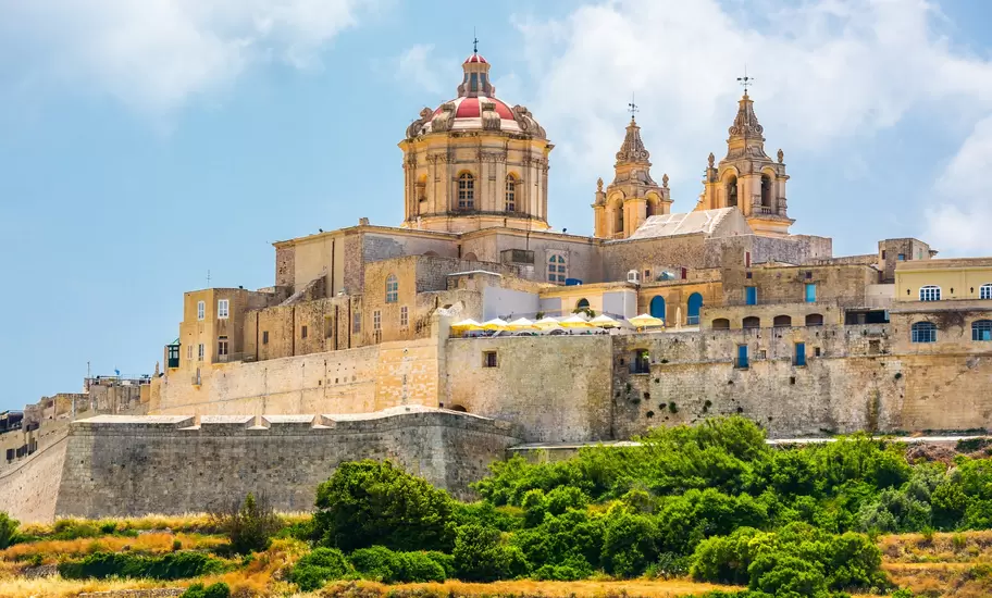 Mdina, Malta: honey-coloured bastion walls topped by the dome and twin bell towers of St Paul's Cathedral, green scrub below