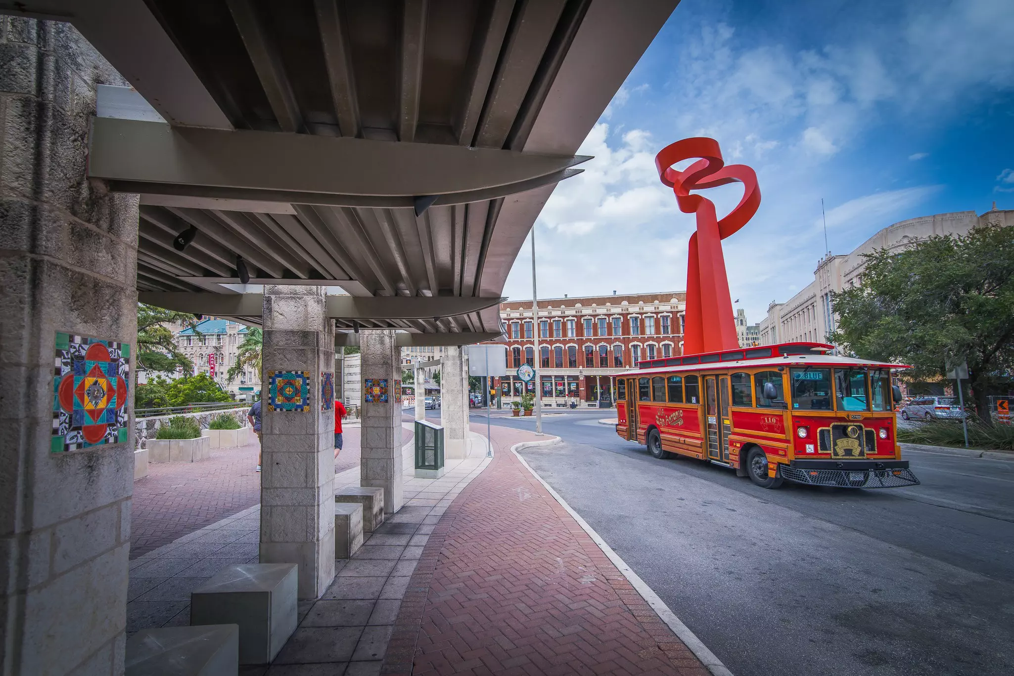 A red tour bus driving past the Torch of Friendship statue in downtown San Antonio, Texas.