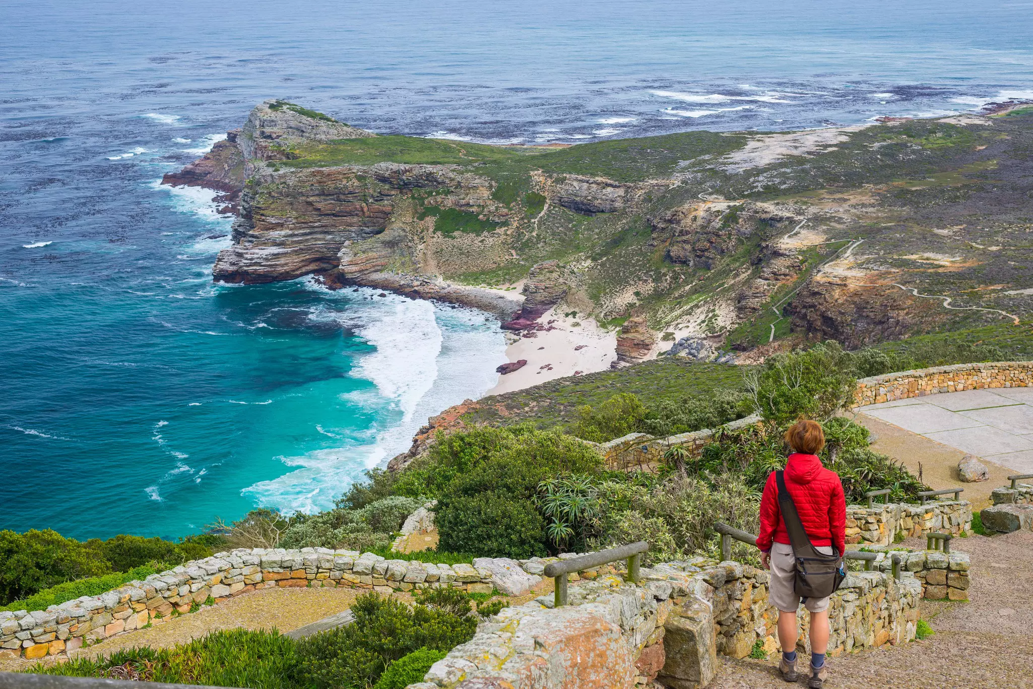 A hiker pauses on a paved coastal trail to look down towards a sandy beach and a peninsula jutting out to sea.