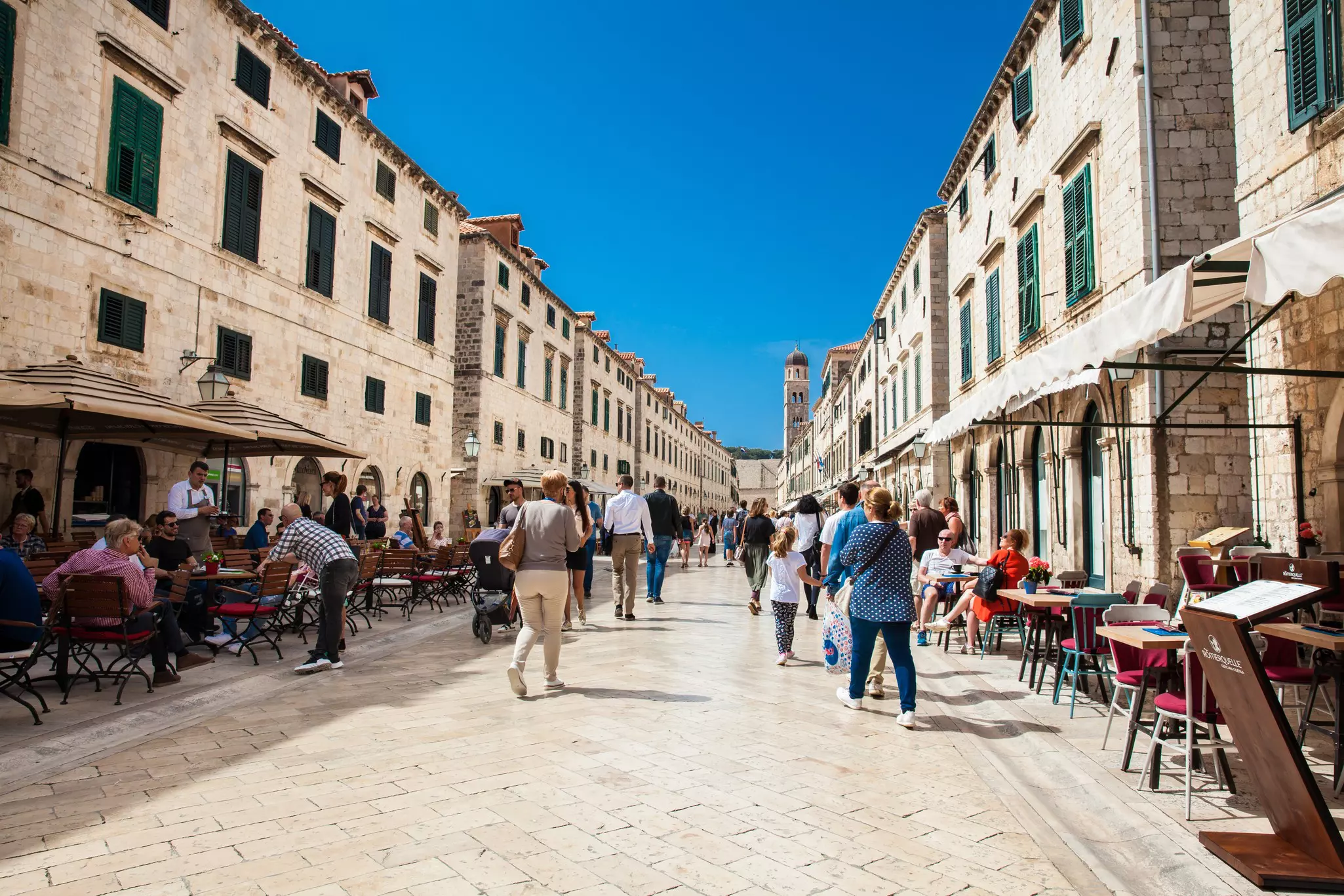 People walk down a long white-stone pathway through the center of an old town. Cafes and restaurants line the sides.