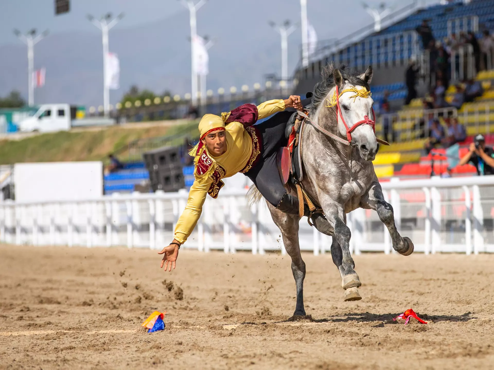 A man on a galloping horse in a hippodrome leans over to grab an object off the dirt track.