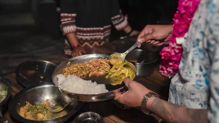 A hand holding a metal dish with rice; they are spooning in additional food items.