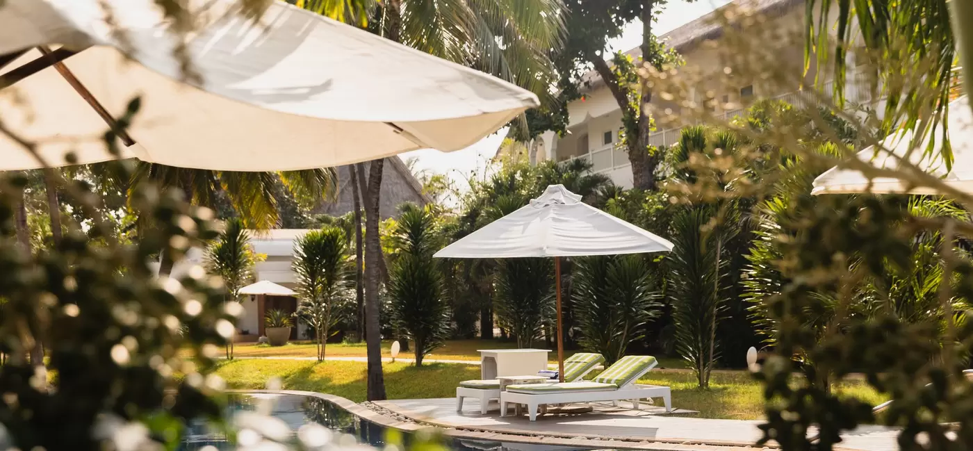 Lounge chairs under a white umbrella by a pool; greenery frames the foreground.
