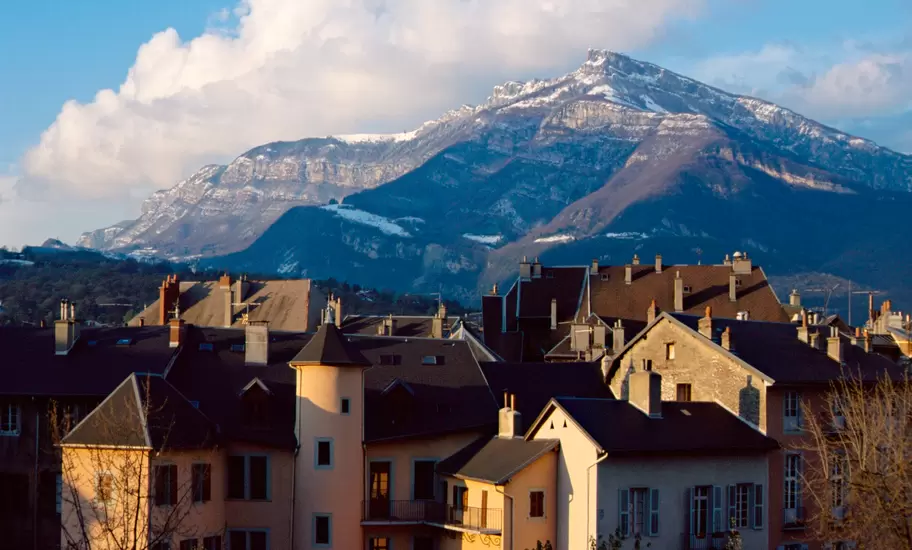 Chambéry rooftops and pastel townhouses with a turret, a snow-dusted limestone mountain and clouds behind