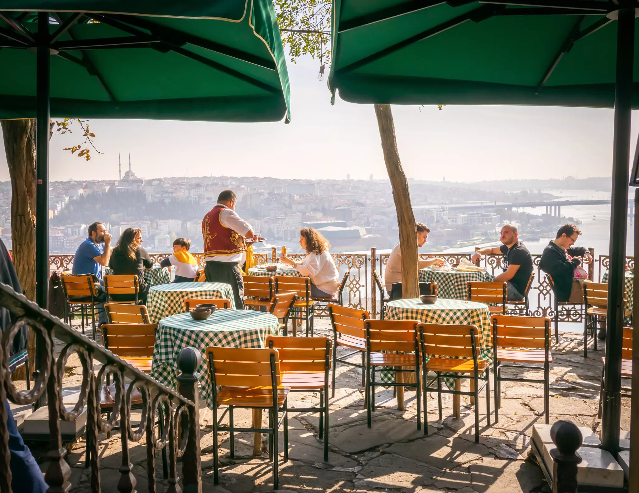 People sitting at an outdoor cafe overlooking a waterway and city buildings in the far distance on a sunny day.