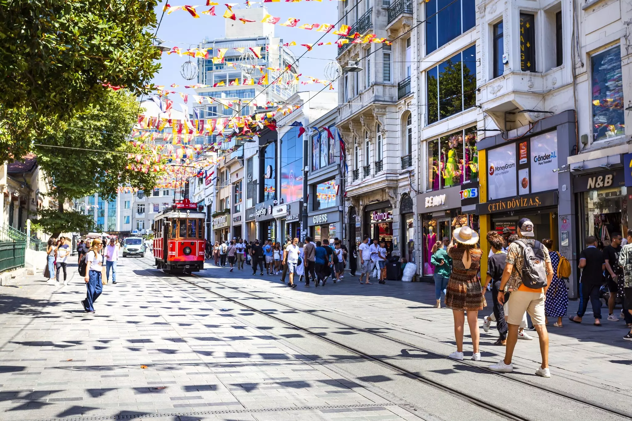 A red tram runs down the center of a shop-filled street with many shoppers browsing and buying.