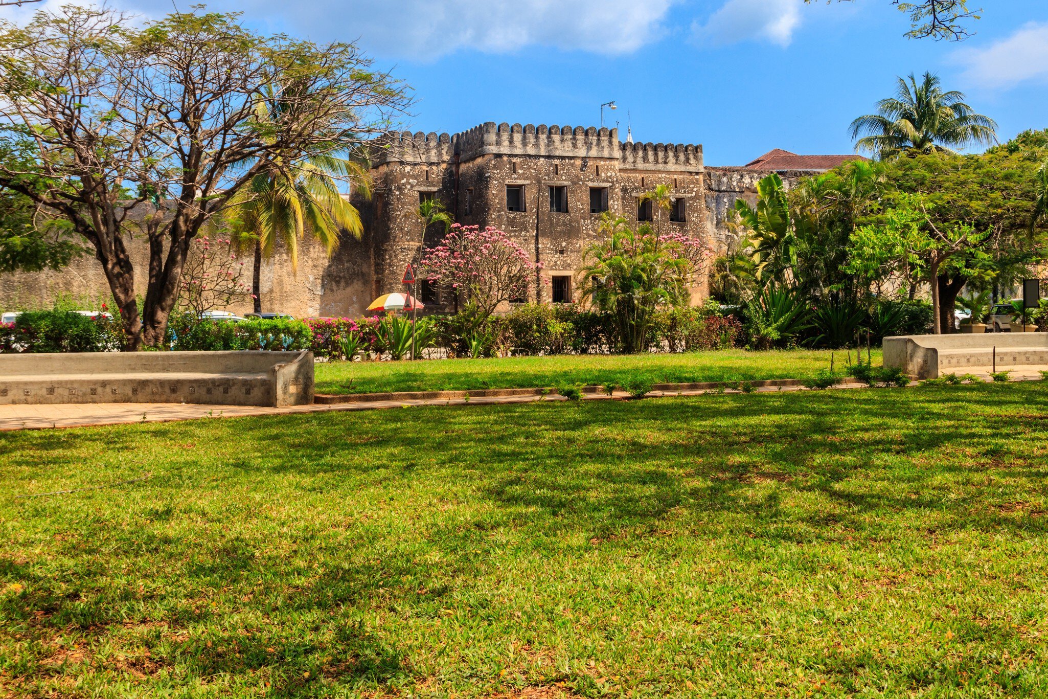 A crenellated coral-stone fortress behind flowering shrubs and palms, seen across a wide lawn with low stone benches.