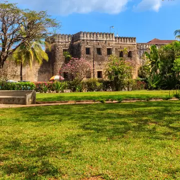 A crenellated coral-stone fortress behind flowering shrubs and palms, seen across a wide lawn with low stone benches.