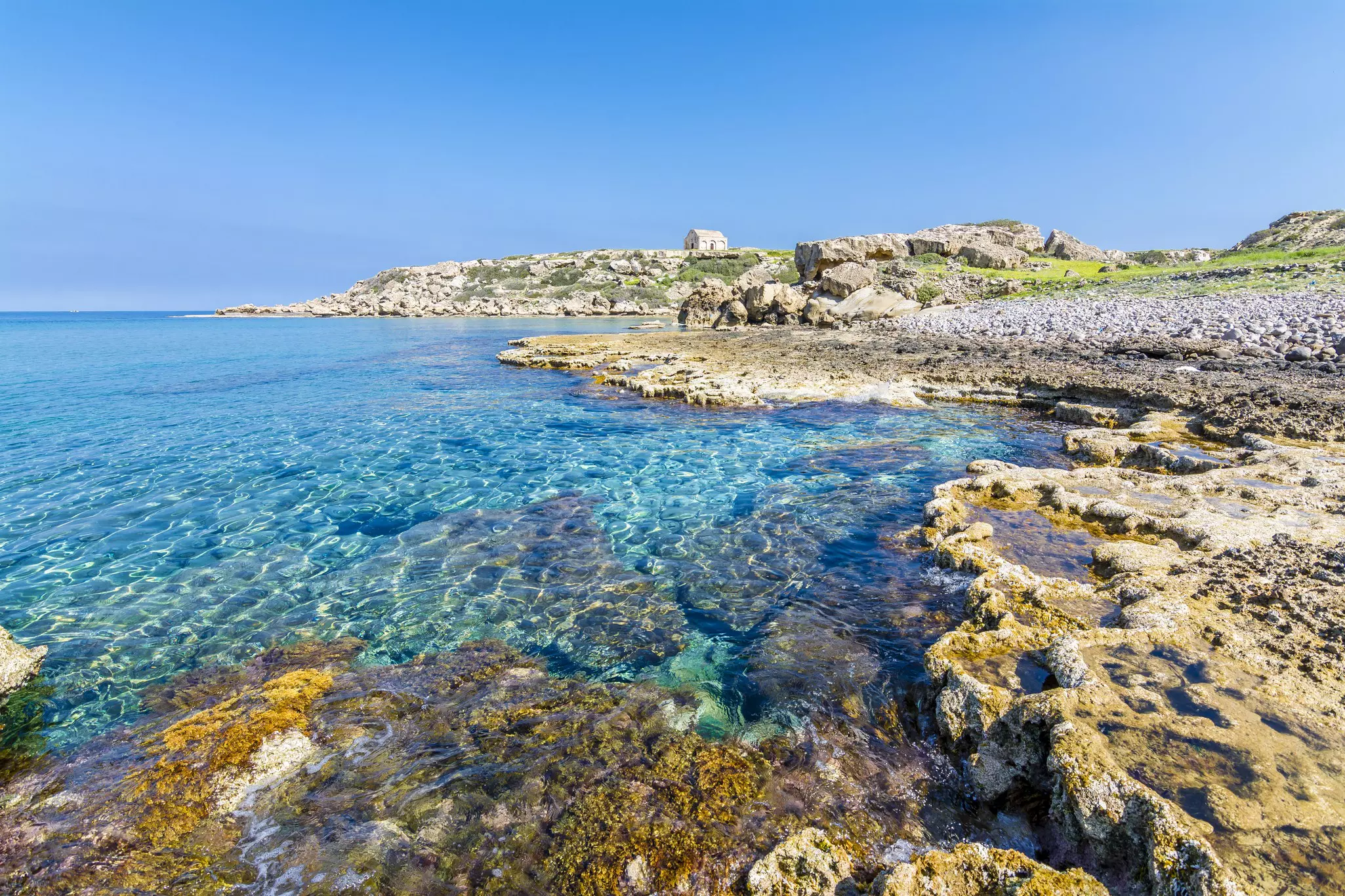 A rocky shoreline with a small stone building in the distance. The sea is clear and very still.