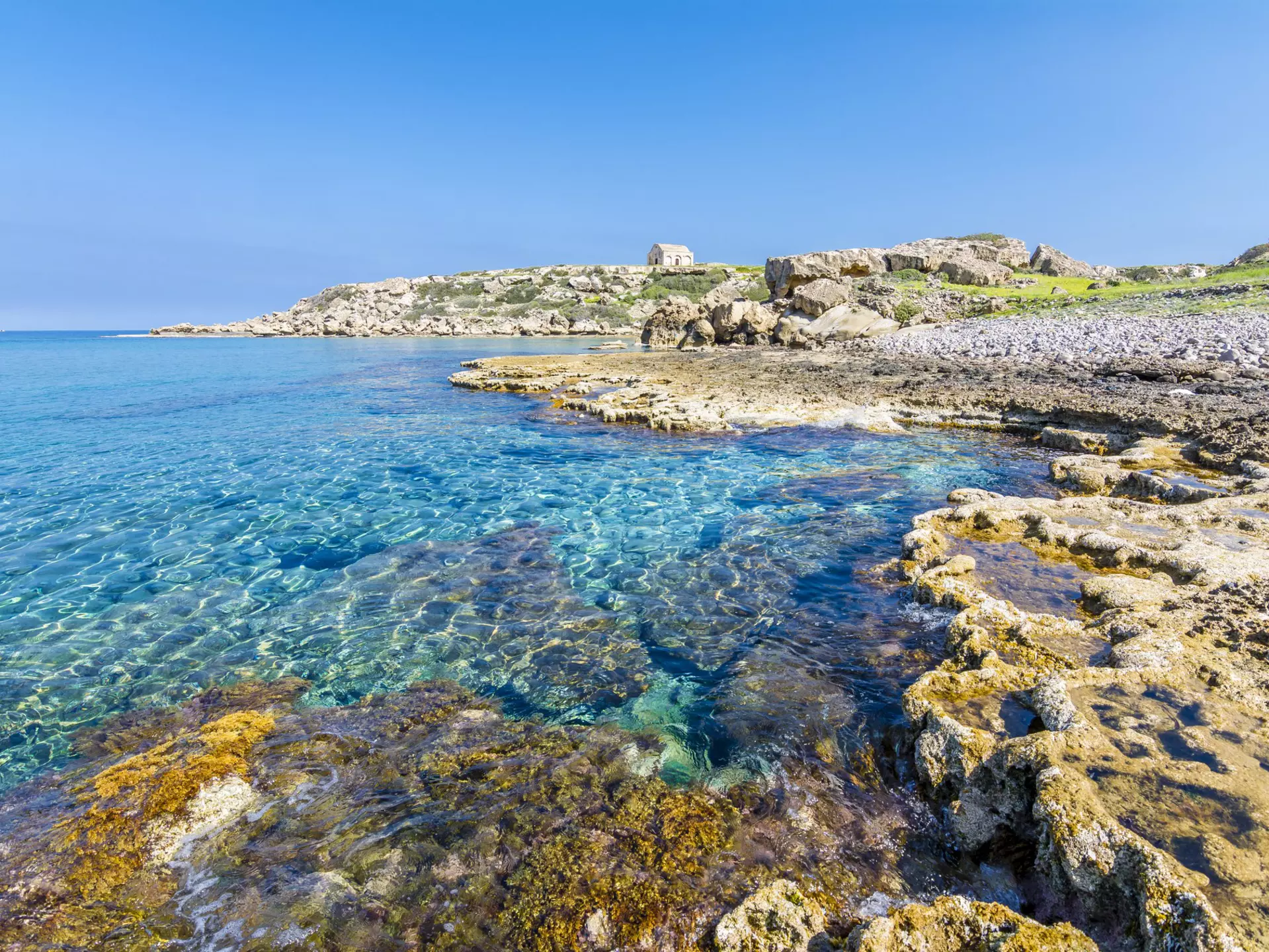 A rocky shoreline with a small stone building in the distance. The sea is clear and very still.