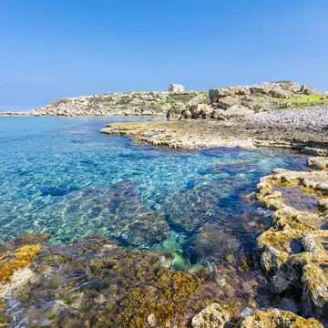 A rocky shoreline with a small stone building in the distance. The sea is clear and very still.