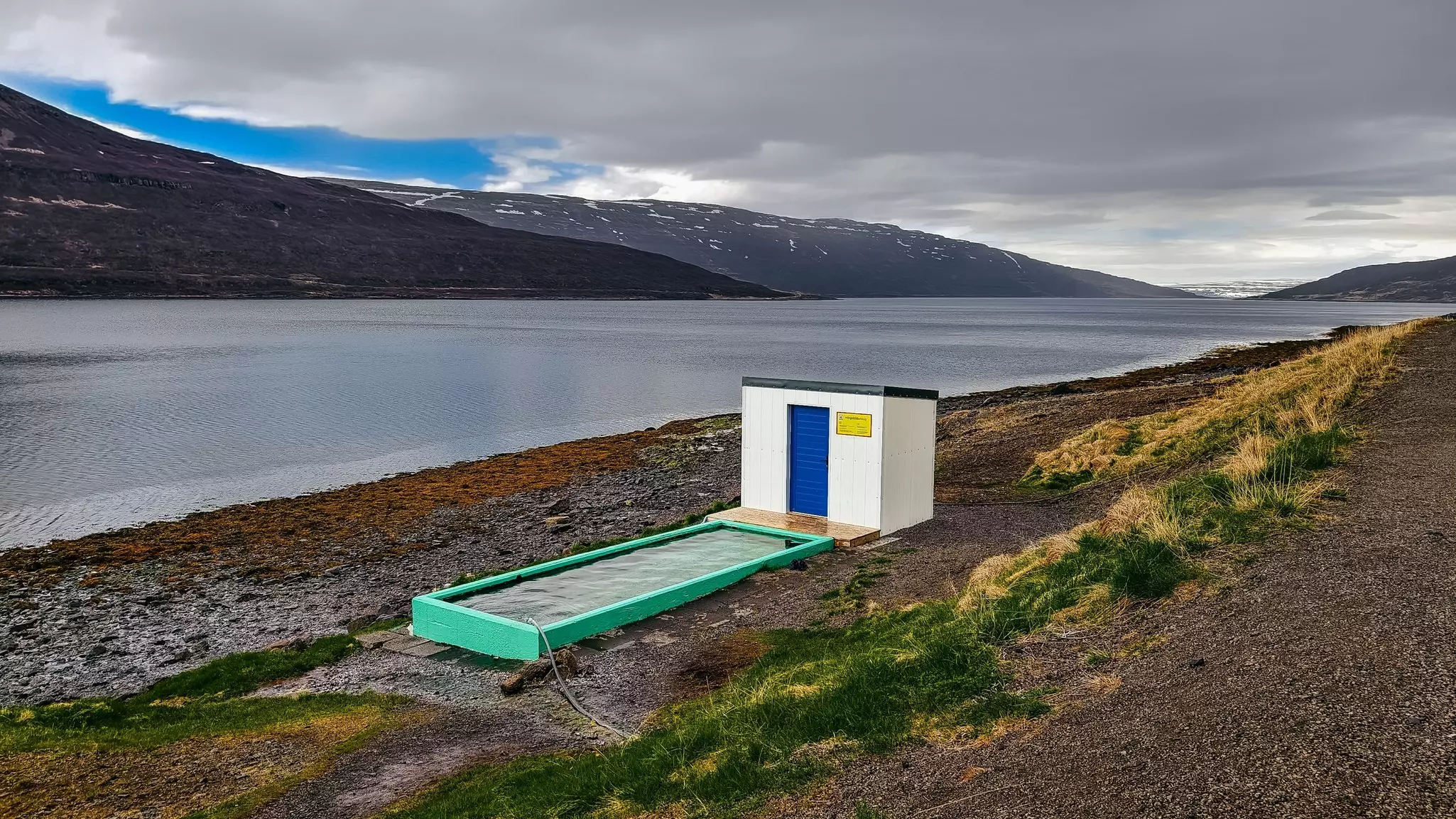 Rectangular hot tub in the sand along a waterway with low mountains and clouds in the distance.