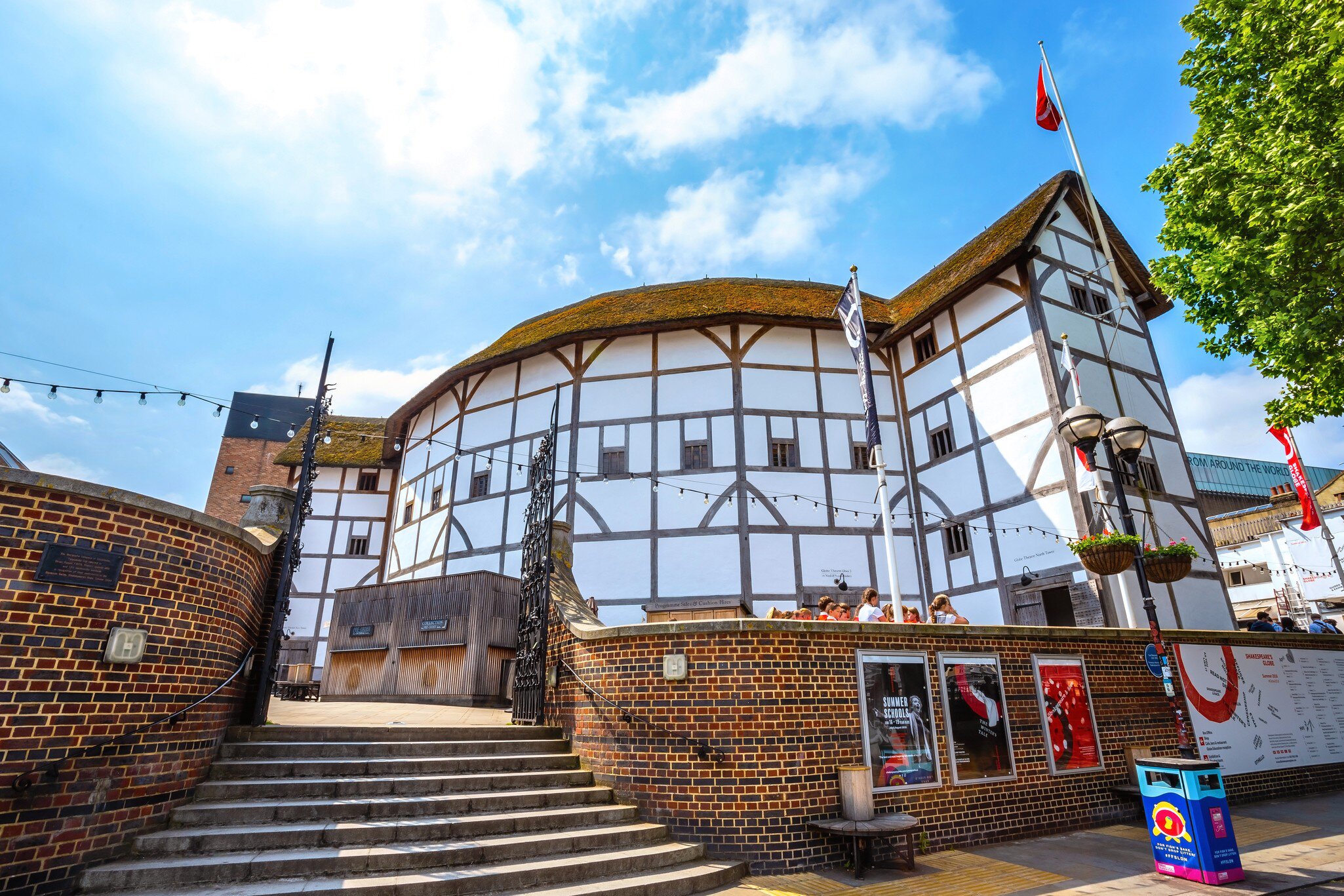 White half-timbered round playhouse under a thatched roof, above a curved brick wall, steps, flags and posters.